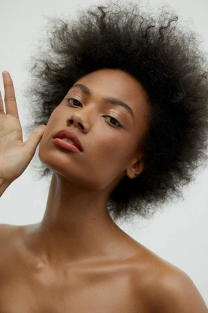 Close-up portrait of an adult woman with dark, curly hair and tan skin. She has her hand up near her face and is indoors with a light background.
