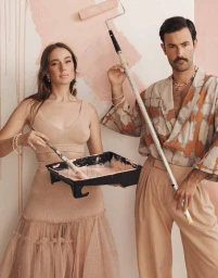 Man and woman painting a wall, holding a paint roller and paint tray in a styled indoor setting.