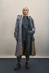 Model with blonde hair wearing a grey trench coat over a matching jumpsuit and dark shoes against a seamless backdrop