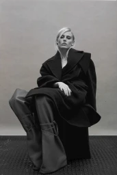 Monochrome portrait of a woman seated in a dark coat and tall boots against a plain studio backdrop