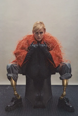 Model with prosthetic legs seated wearing an orange feather coat and black leather gloves