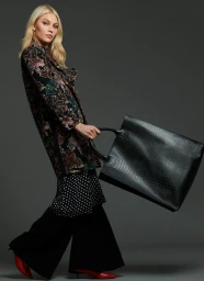 A blonde model wearing a floral patterned coat and black trousers holds a large black tote bag against a grey seamless background.