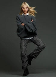 Blonde woman with arms crossed wearing a grey sweater, white collared shirt, and plaid trousers against a grey backdrop