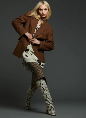 Model with blonde hair wearing a brown blazer, patterned top, and snakeskin boots against a dark grey seamless studio backdrop.