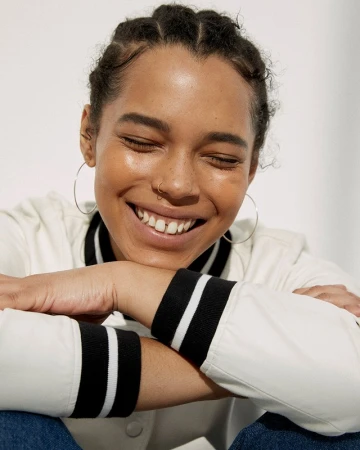 Smiling woman with braided hair and hoop earrings wearing a white jacket resting her chin on crossed arms against a plain white background.