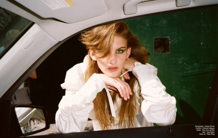 A young woman with long brown hair and green eye makeup leans through a car window, wearing a white blouse.