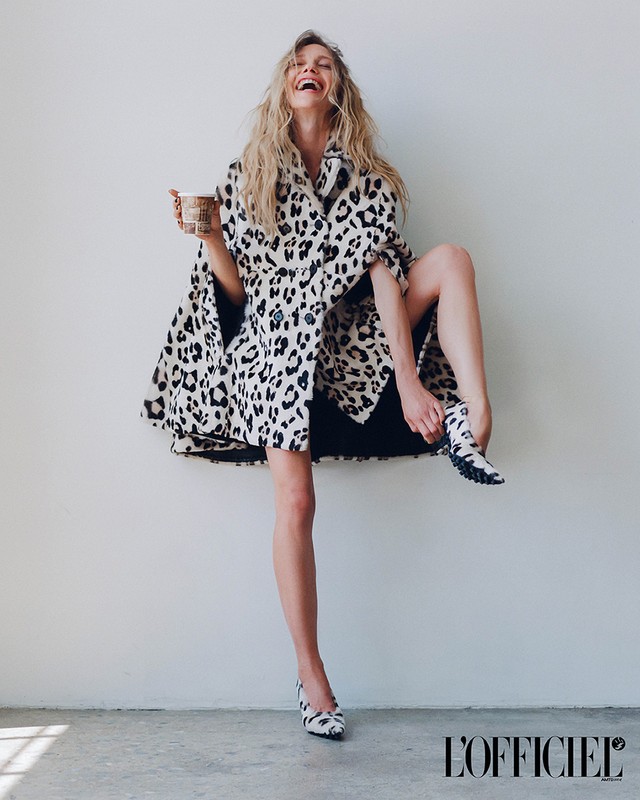A blonde woman in a black and white animal print coat laughs while standing in front of a wall, holding a coffee cup.