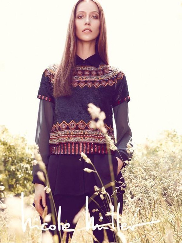 Fashion photo of a young adult woman in an ornate top, standing in a field of tall grasses, with signature text overlay.