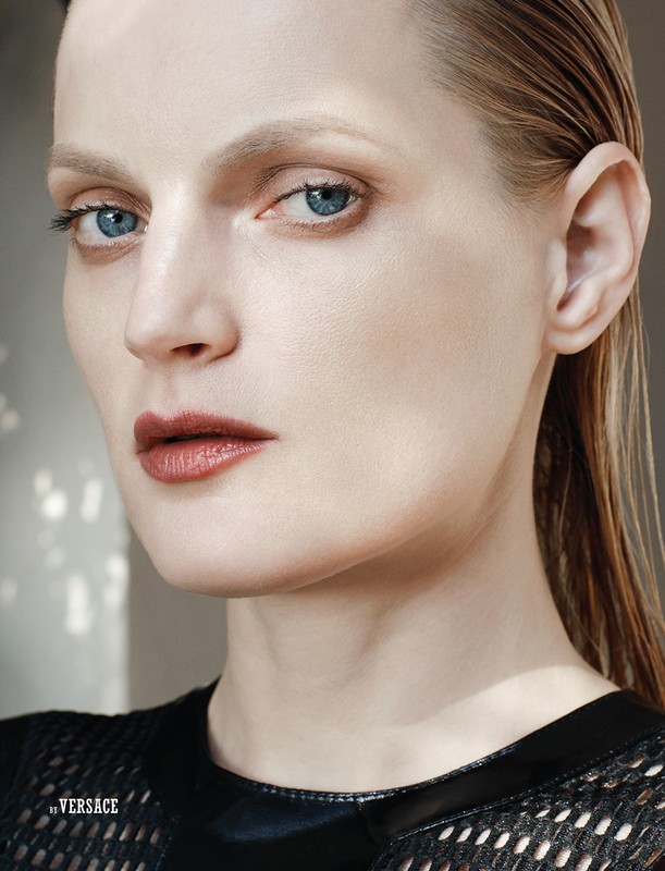 Close-up portrait of an adult woman with blonde hair and blue eyes, wearing a black mesh top, set against a neutral background with dappled lighting.