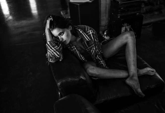 Monochrome portrait of a woman in a plaid shirt reclining on a leather sofa, looking at the camera in an indoor setting.
