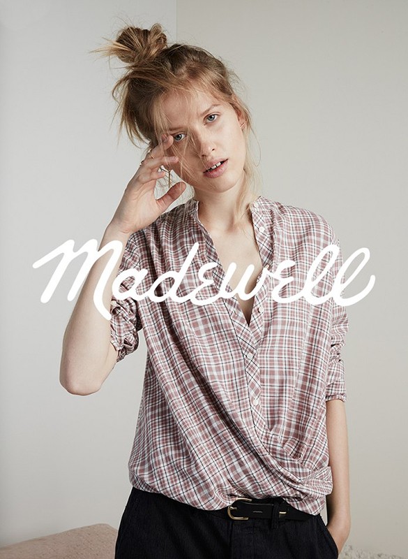 A woman poses indoors wearing a plaid collared shirt and dark pants, with the brand name 'Madewell' in large white font overlayed.