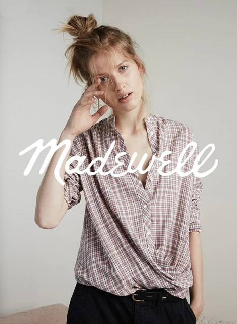 A woman poses indoors wearing a plaid collared shirt and dark pants, with the brand name 'Madewell' in large white font overlayed.