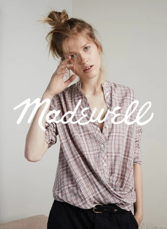 A woman poses indoors wearing a plaid collared shirt and dark pants, with the brand name 'Madewell' in large white font overlayed.