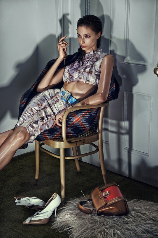 A woman in patterned crop top and skirt sitting on a chair with handbag and shoes on the floor in a simple indoor setting.