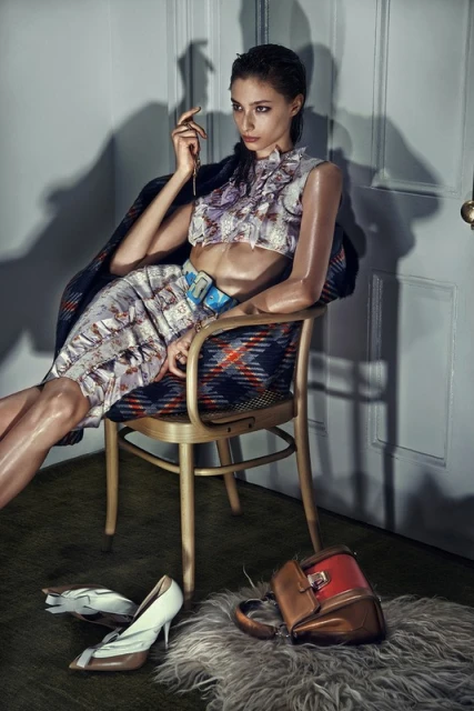 A woman in patterned crop top and skirt sitting on a chair with handbag and shoes on the floor in a simple indoor setting.