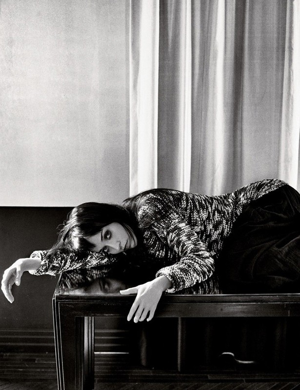 Monochrome portrait of a woman with dark hair lying across a dark wooden table in front of vertically draped curtains.