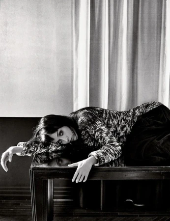 Monochrome portrait of a woman with dark hair lying across a dark wooden table in front of vertically draped curtains.