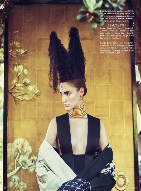 Model poses in avant-garde fashion, featuring a black dress and ornate feathered headdress, against a golden background with foliage.