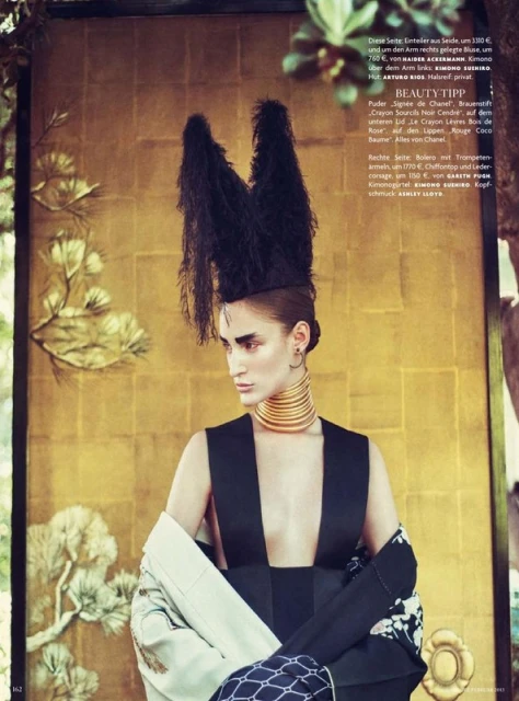 Model poses in avant-garde fashion, featuring a black dress and ornate feathered headdress, against a golden background with foliage.