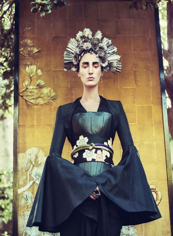 Fashionable woman in black couture with ornate floral headdress poses against a gold-painted panel in an outdoor garden setting.