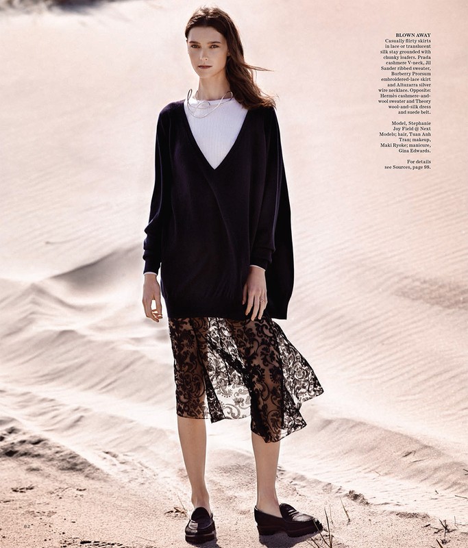 Fashion editorial shot of a woman wearing a dark, sheer, lace skirt with a black top and loafers, posed on a sand dune.