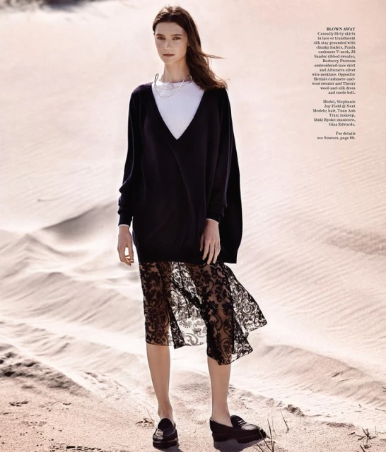 Fashion editorial shot of a woman wearing a dark, sheer, lace skirt with a black top and loafers, posed on a sand dune.