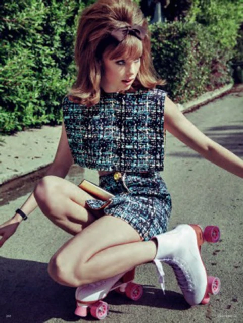 A woman on roller skates is wearing a two-piece turquoise tweed outfit with a brown hair bow.  She is outdoors surrounded by green foliage.