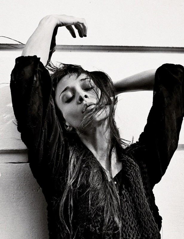 Black and white portrait of a woman with dark hair styling her hair with her hands raised, wearing a textured black top.