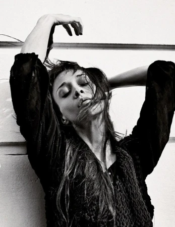 Black and white portrait of a woman with dark hair styling her hair with her hands raised, wearing a textured black top.