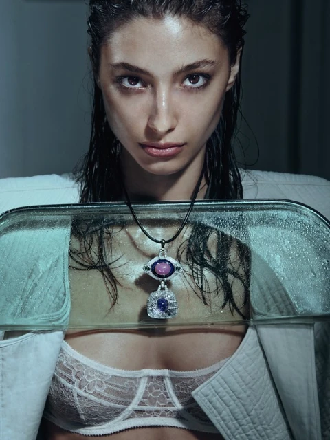 Portrait of a woman with wet hair wearing a white coat, posing with a reflective object and a gemstone necklace in a studio.