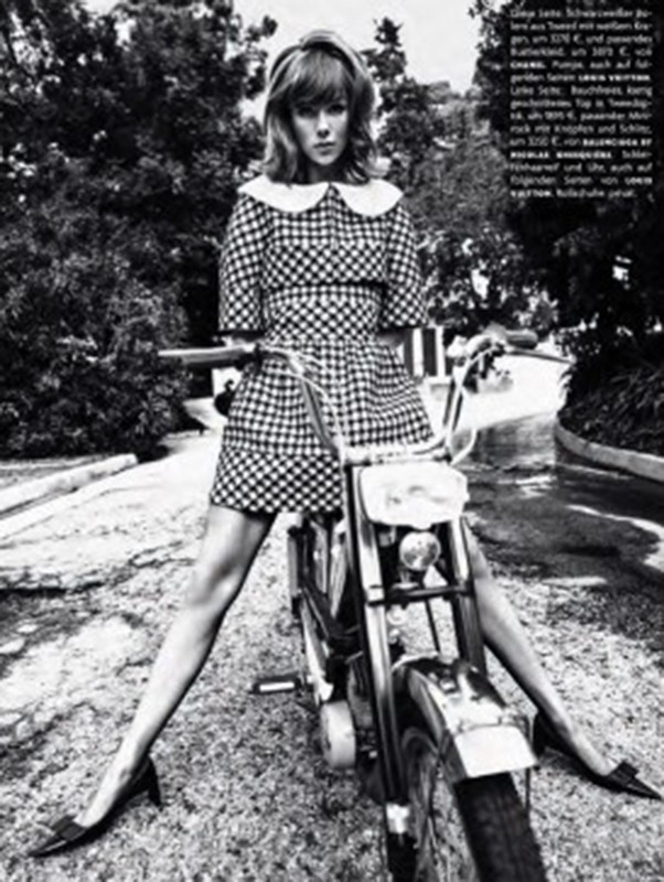 A woman in a checkered dress sits astride a motorcycle in a black and white photo, framed by trees and a paved road. 
