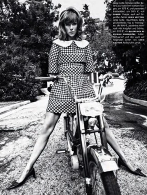 A woman in a checkered dress sits astride a motorcycle in a black and white photo, framed by trees and a paved road. 