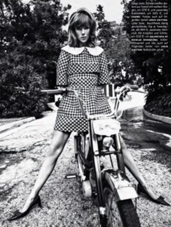 A woman in a checkered dress sits astride a motorcycle in a black and white photo, framed by trees and a paved road. 