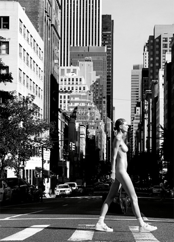 Model walking across a city street crosswalk wearing white sneakers, captured in a monochrome urban scene.