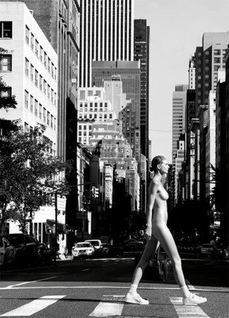 Model walking across a city street crosswalk wearing white sneakers, captured in a monochrome urban scene.