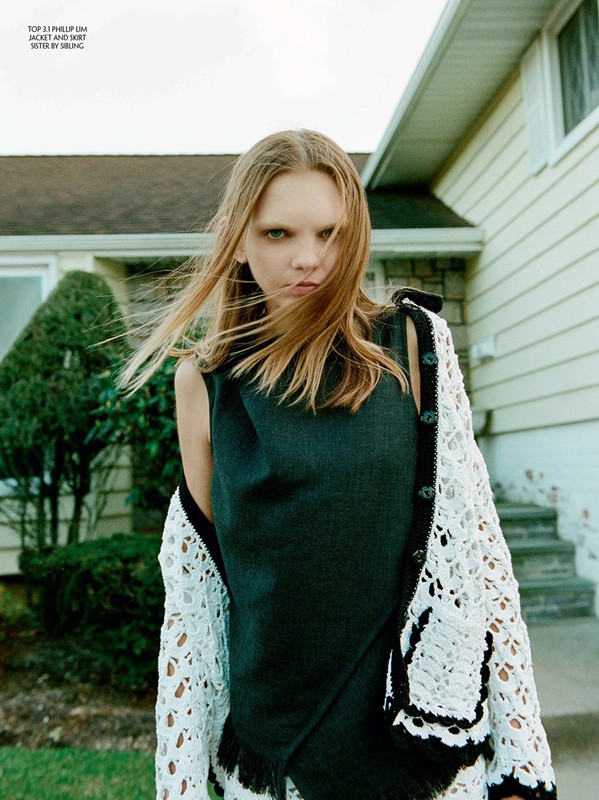 A fair-skinned fashion model with light brown hair in a gray tank top and white crochet cardigan poses outside a suburban home.