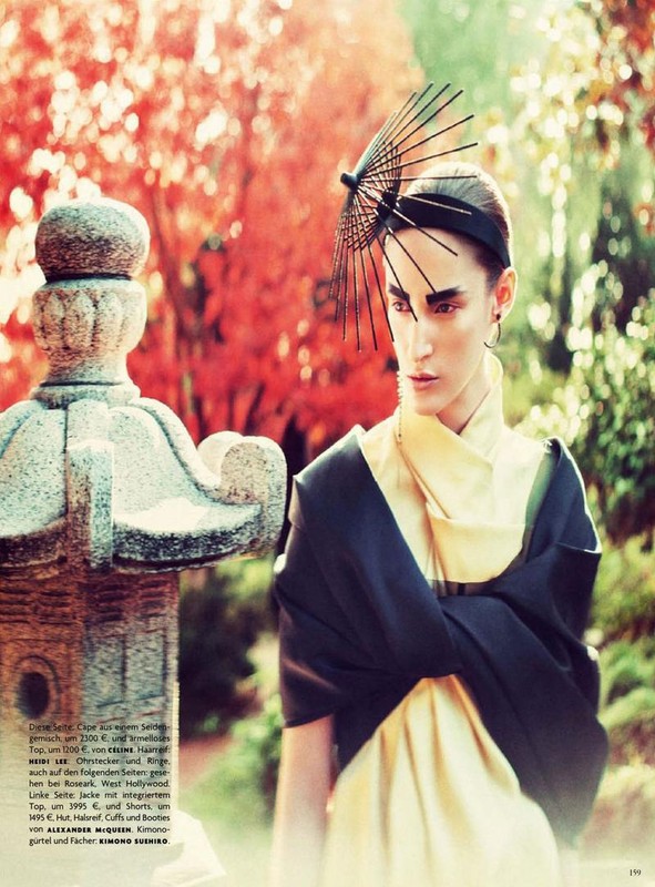 Woman in kimono-style dress and headpiece poses in garden with autumn foliage and stone lantern. Fashion editorial image.