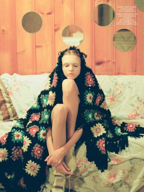 Young girl wrapped in a floral blanket sits on a bed. The room has a light orange wall with round mirrors as decoration.