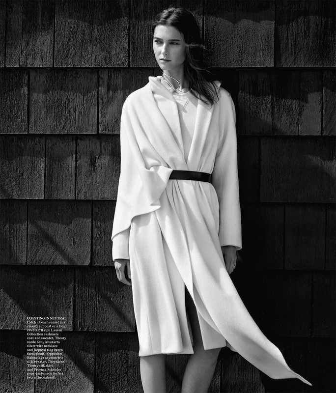 Monochrome studio shot of a Caucasian woman with dark hair, wearing a white coat with a black belt, posing against a wood wall.