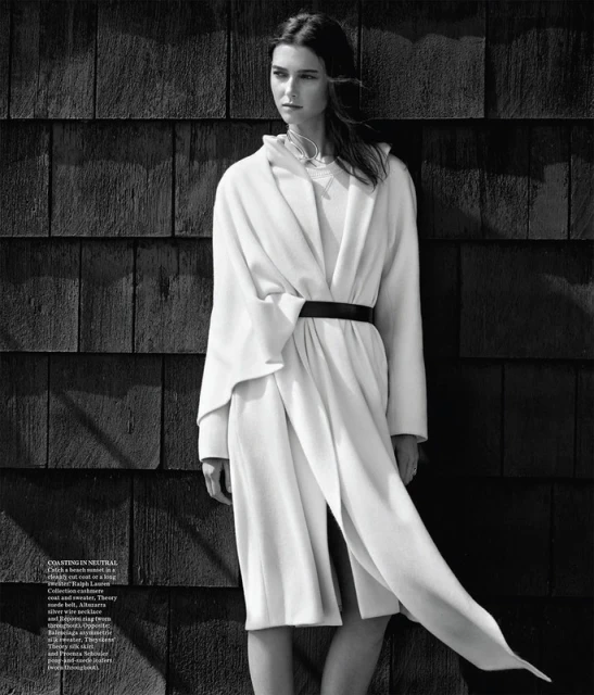 Monochrome studio shot of a Caucasian woman with dark hair, wearing a white coat with a black belt, posing against a wood wall.