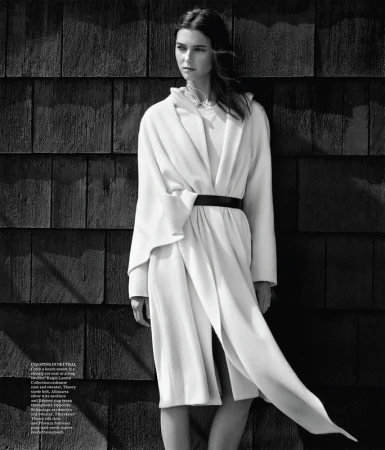 Monochrome studio shot of a Caucasian woman with dark hair, wearing a white coat with a black belt, posing against a wood wall.