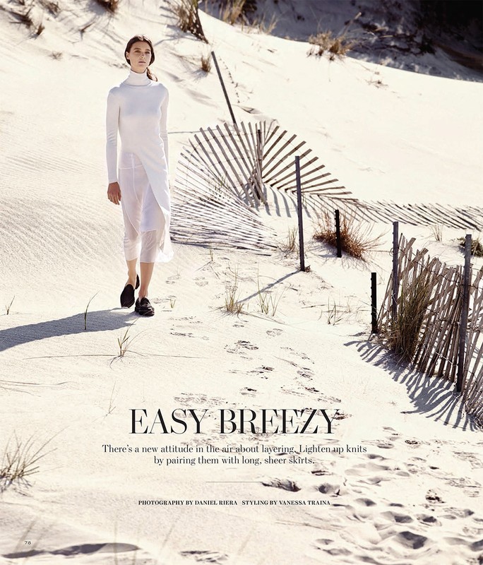 A woman in a long white dress walking on a sand dune with a broken fence, captured in bright daylight, showcasing fashion and nature.