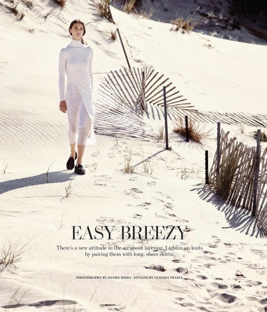 A woman in a long white dress walking on a sand dune with a broken fence, captured in bright daylight, showcasing fashion and nature.