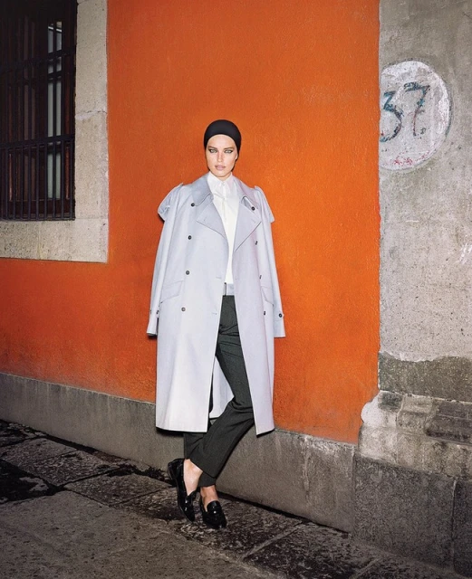 A woman in a light gray trench coat stands in front of an orange wall on a city street, styled in a turban and dark slacks and loafers.