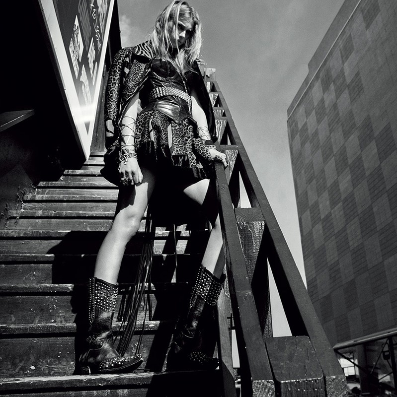 A woman poses on wooden stairs outdoors, wearing a jacket, dress, and boots with studs against an urban background with a tall building.