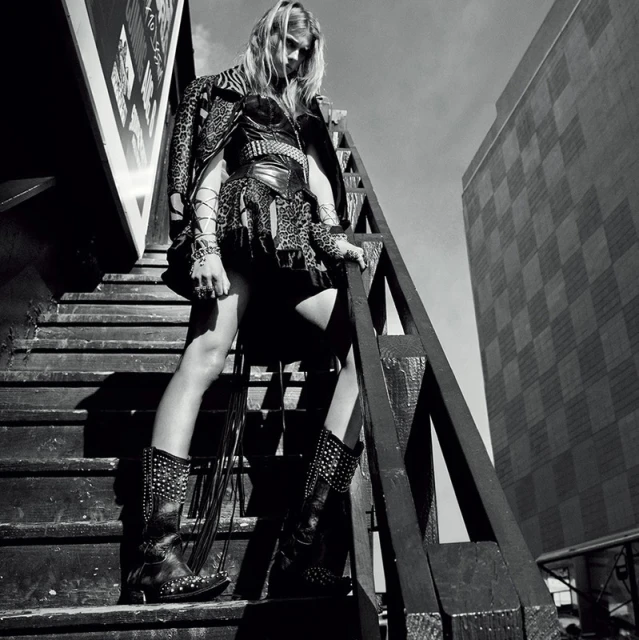A woman poses on wooden stairs outdoors, wearing a jacket, dress, and boots with studs against an urban background with a tall building.