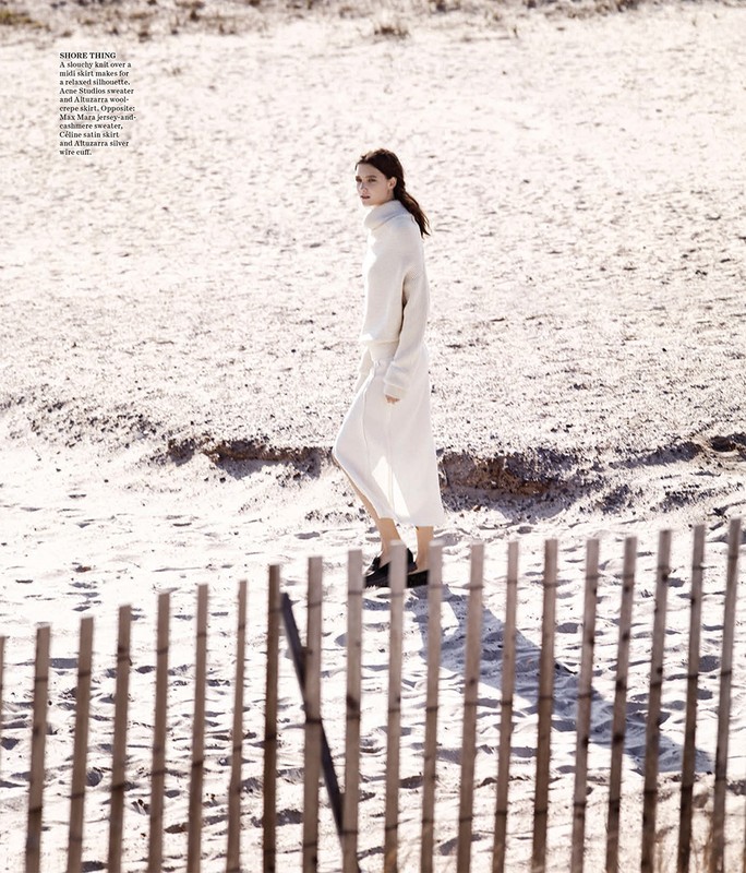 A woman in a white sweater and long skirt walks on a sandy beach near a light brown wooden fence under bright, diffused sunlight.