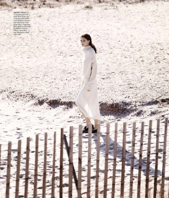 A woman in a white sweater and long skirt walks on a sandy beach near a light brown wooden fence under bright, diffused sunlight.