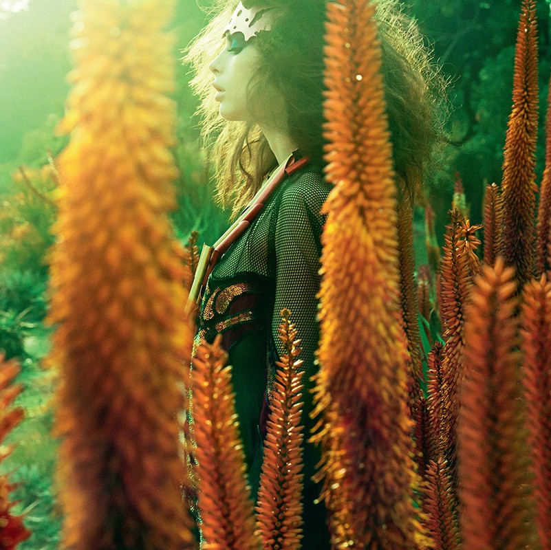 A woman with a decorative headpiece poses in a garden amongst orange flowering plants in a natural, outdoor setting with soft lighting.