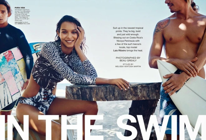Fashion editorial shot featuring a model in a patterned swimsuit, seated on a wooden surface, with two surfers holding boards in the background.